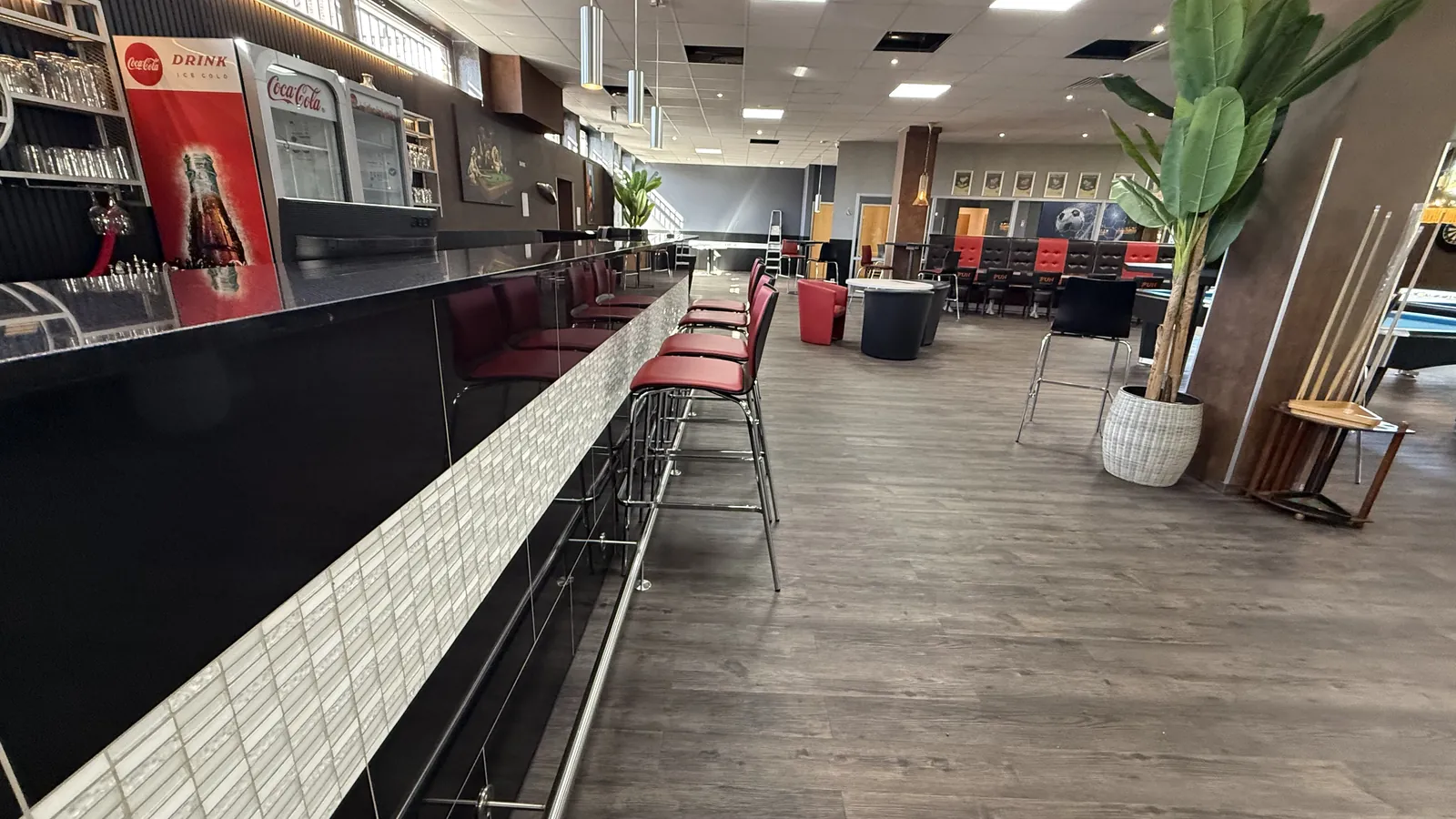 Lange Bartheke mit weißer Mosaikfront und roten Barhockern in der Sportsbar Voerde, dahinter der Saal mit Sitzgruppen und Billardtischen